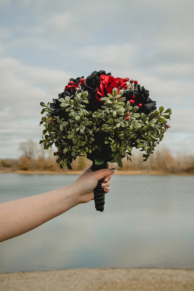 bride's arm extending in from out of frame to showcase red and black rose bouquet with accent greenery