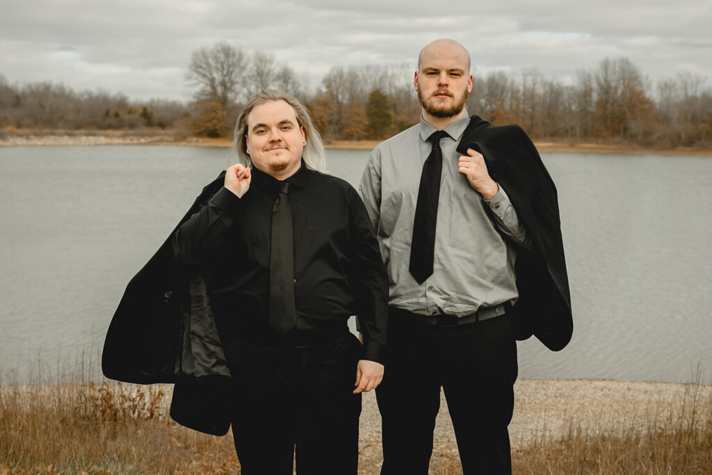 young groom in all-black suite and his best man in a gray shirt and black tie smirk at the camera while holding their suit jackets over their shoulders