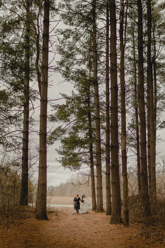 Mother of the bride and bride walking down an aisle clearing dwarfed by a grove of tall pine trees