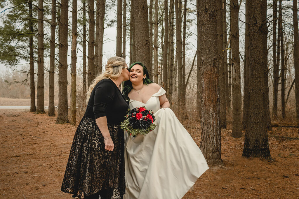 mother of the bride gives her daughter.a kiss on the check as she smiles in front a forest