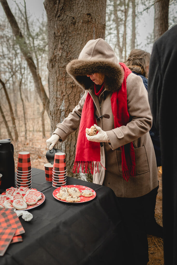 a women in a heavy winter coat poars hot chocolate outside at a table while holding a cookie