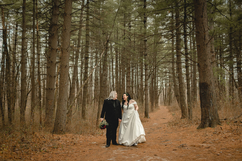 groom holding bride's bouquet as they laugh towards each other while walking through a forest