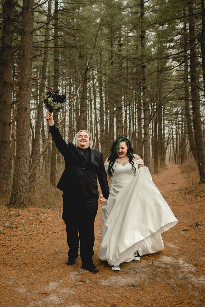 groom smiles toward the camera and hold bride's bouquet up triumphantly as he walks with his bride through a forest
