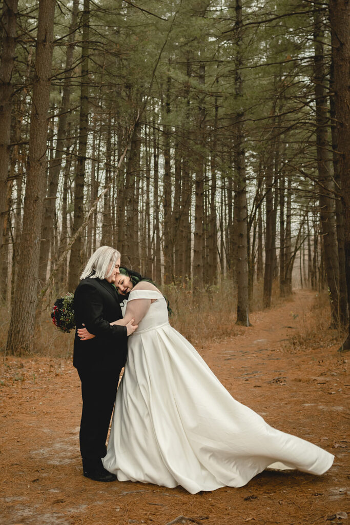 far wide shot of a young bride in white and groom in black hugging in a beautiful pine forest 