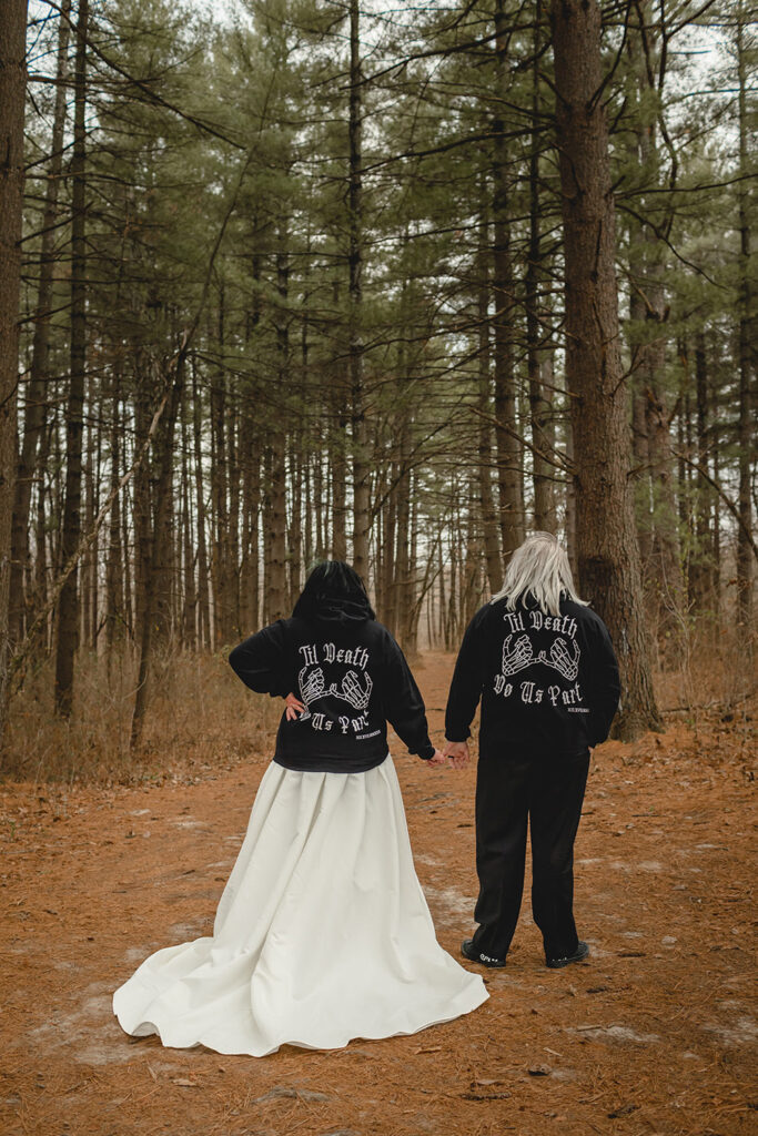 bride and groom with matching black hoodies over their wedding attire, holding hands while facing away from camera in a pine tree forest