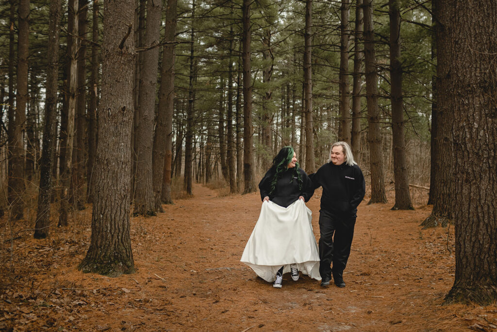 bride and groom wearing matching black hoodies over their wedding attire, smiling towards each other as they walk between tall pine trees