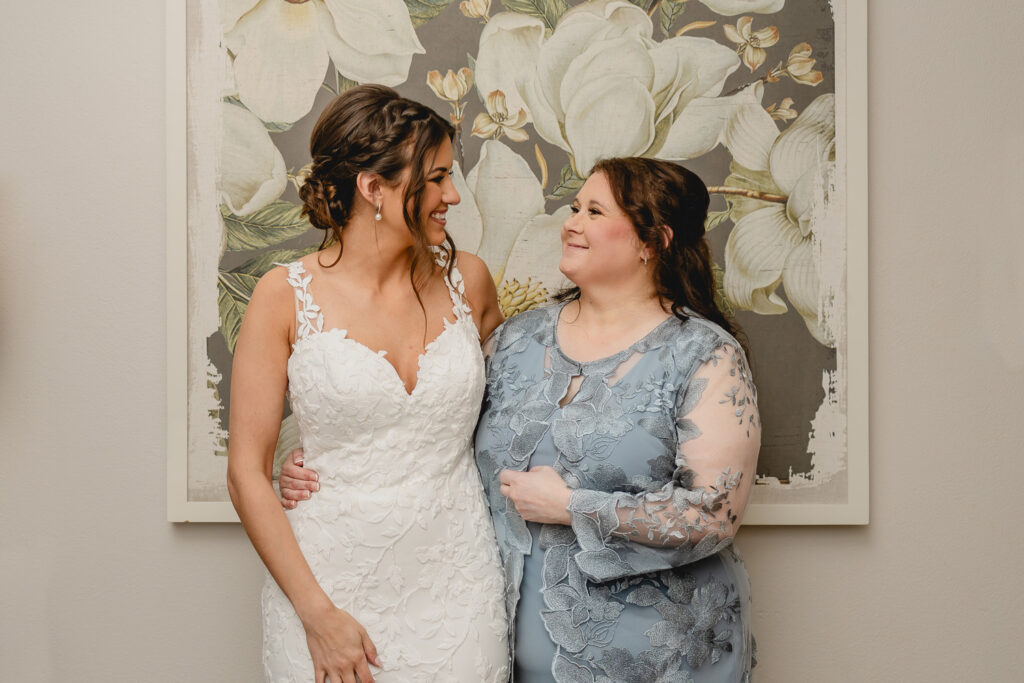 A bride and her mother posing in front of a floral painting smiling at each other