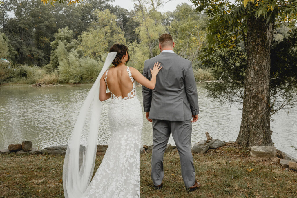 Bride tapping on grooms shoulder during a first look in front of a lake. 