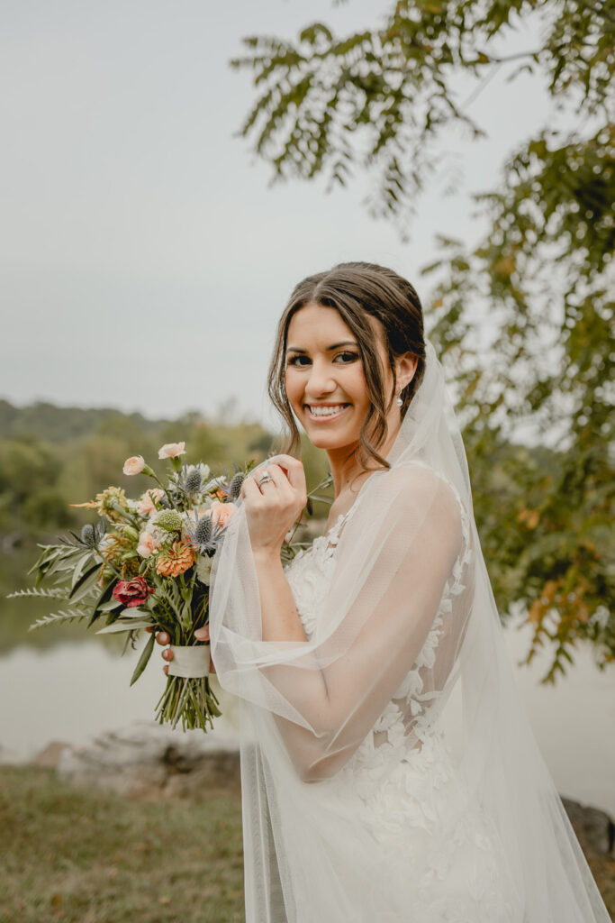 Bride with vail and flowers poses in front of a lake