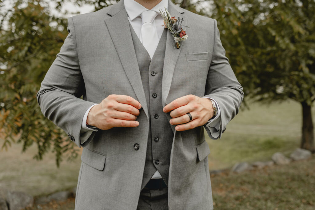 Groom buttoning his suit jacket with close up of wedding ring.