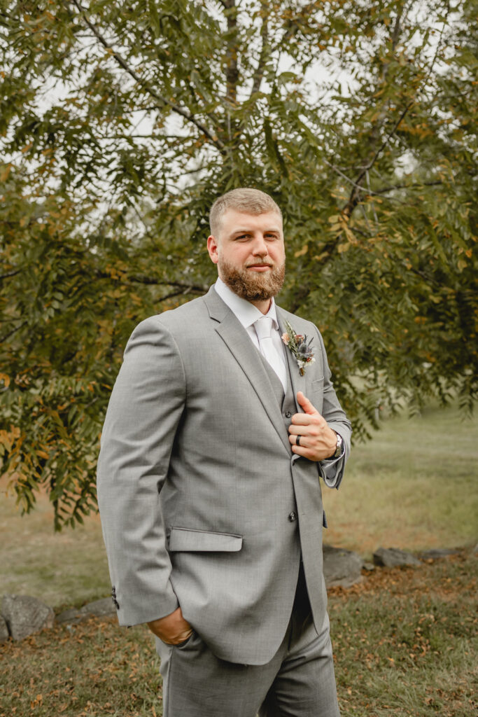 Groom posing in grey suit in front of a tree.