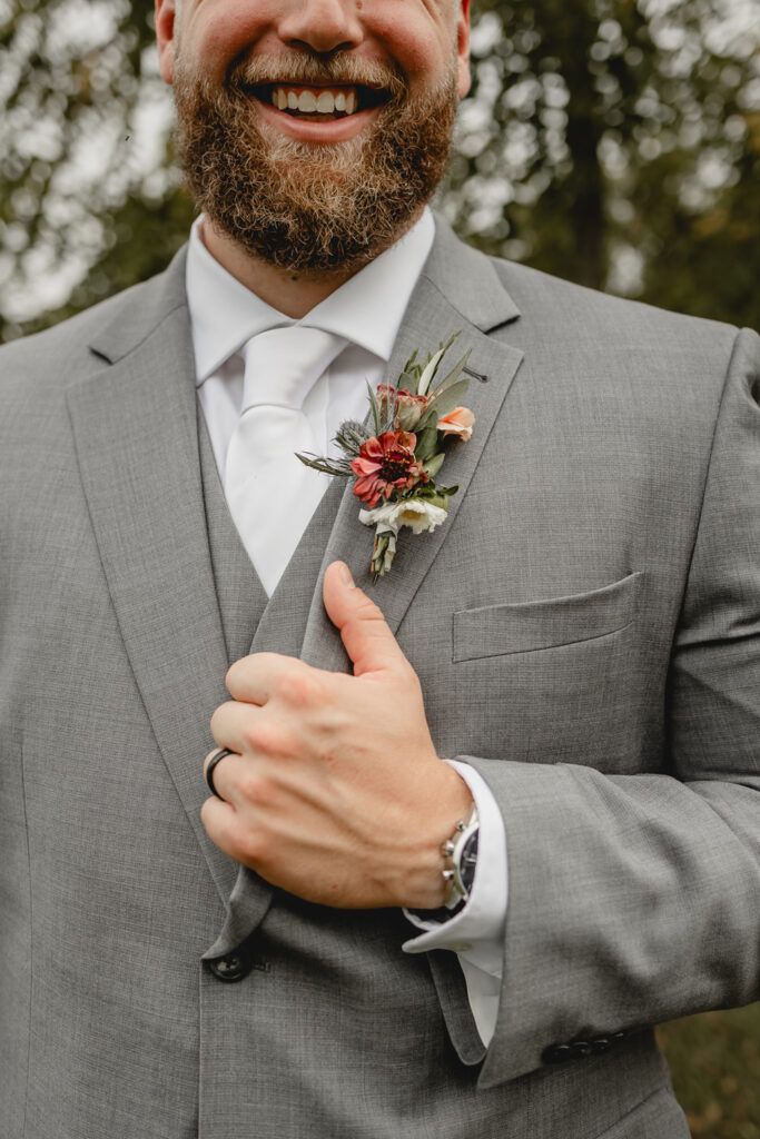 Close up photo of groom smiling and holding his jacket with flower on his lapel.