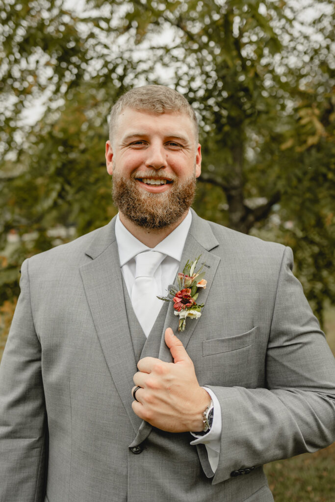 Groom in grey suit poses in front of a tree.