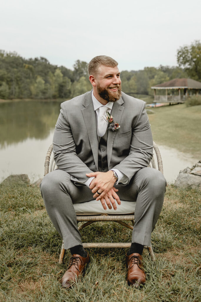 Groom in grey tux sitting on a chair in front of lost hill lake.