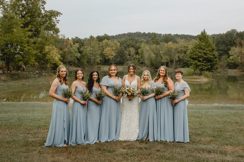 Bridal party posing in blue dresses in front of a lake.
