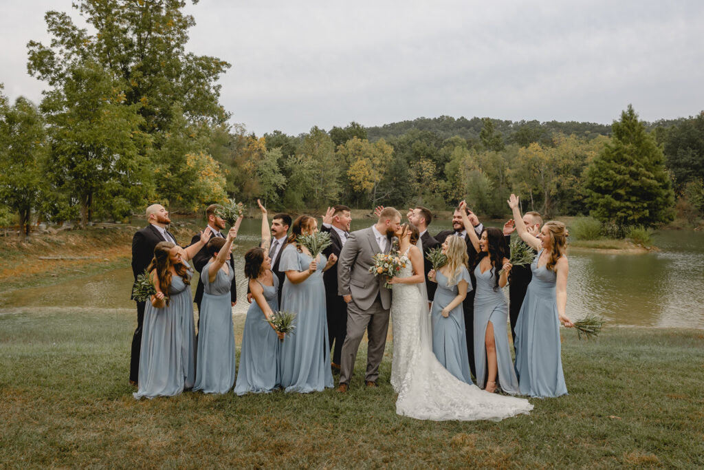 Wedding party celebrates in posed photo against lake backdrop. 