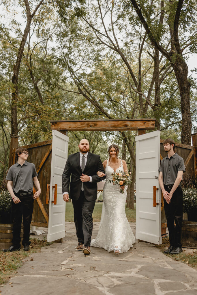 Bride and her brother walking down the aisle through 2 white wooden doors at outdoor ceremony.