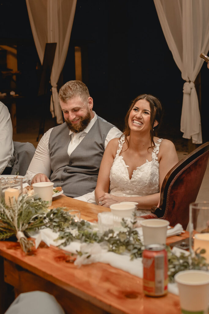 Bride and groom smiling while enjoying brides maid speech seated at table.