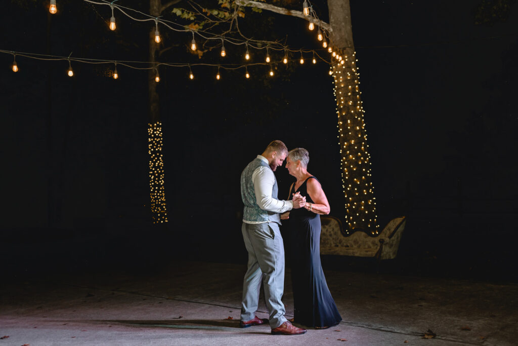 Groom and his mother sharing a dance at wedding reception with string lights hanging above.