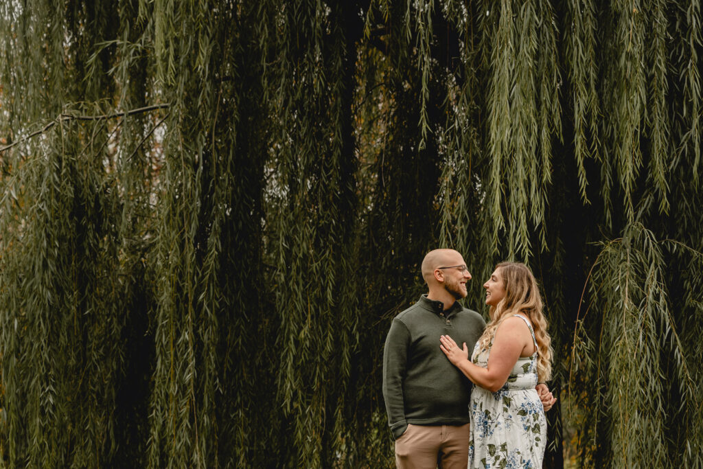 waist up shot of a young couple turned togethers with a large green willow tree fills the background