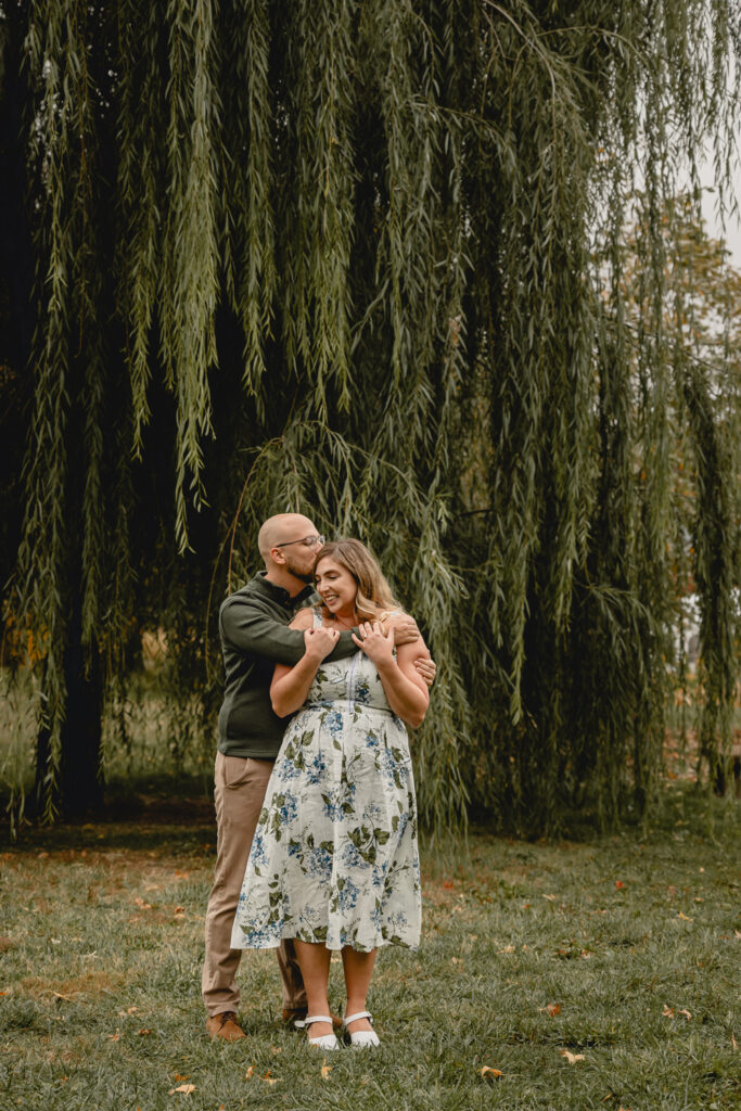 young woman gleefully holds the arms of her boyfriend as he hugs her from behind and kisses her head in front of a giant willow tree