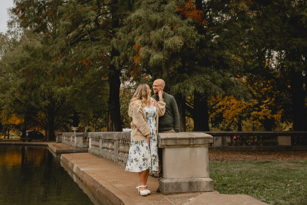 young women in white floral dress and cream cardigan looks back and touches the cheek of her boyfriend as they stand along a stone wall on the edge of a pond