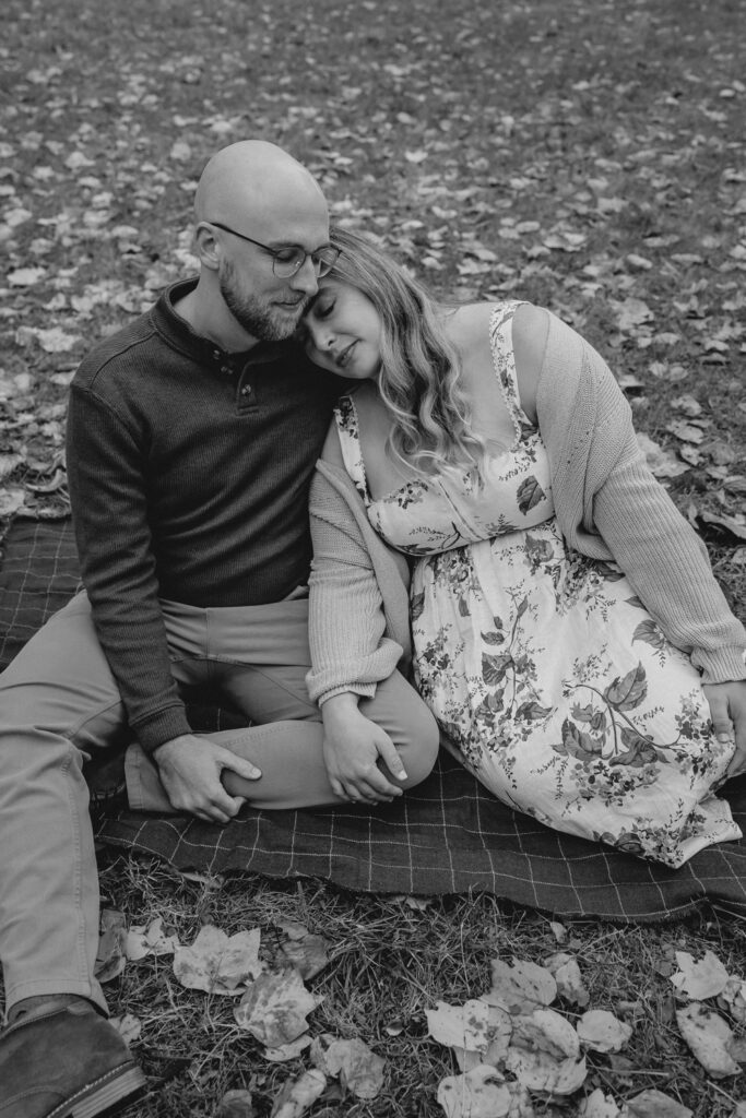moody black and white shot of couple sitting on the grass leaning against each other with eyes closed and surrounded by fallen leaves