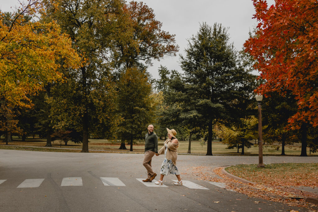 side shot of a couple holding hands and looking lovingly at each other as they cross a crosswalk in a fall park