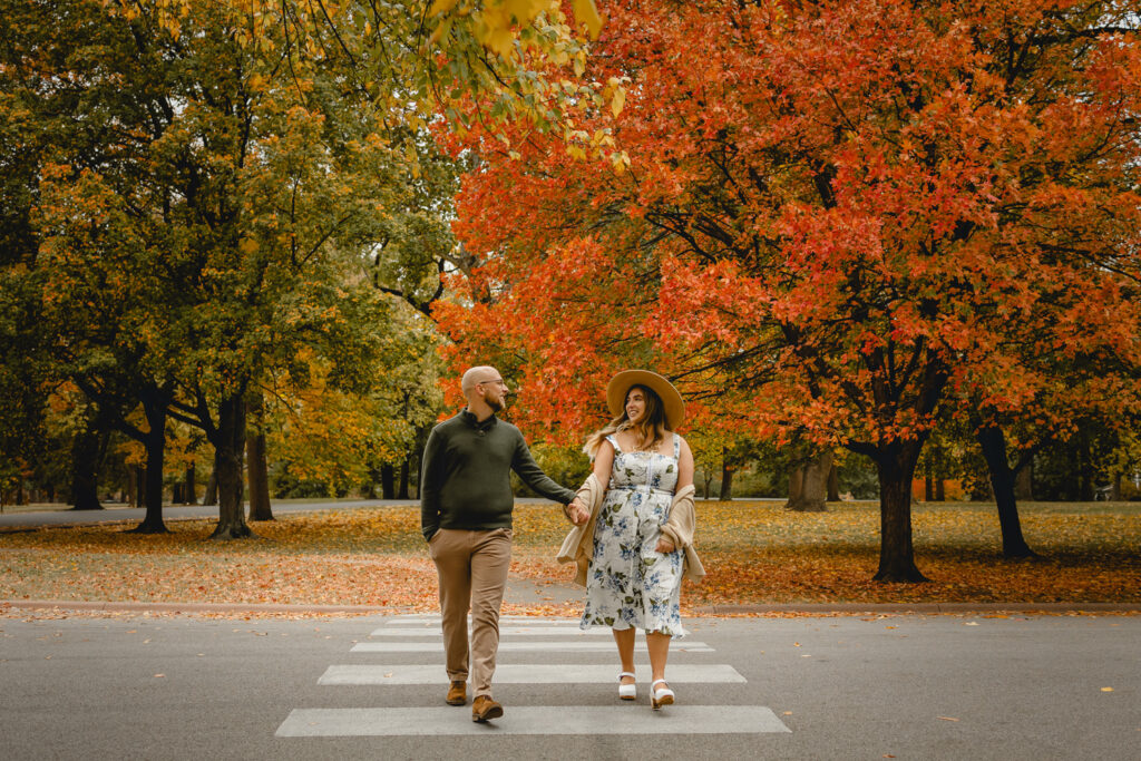 front shot of a couple holding hands and looking lovingly at each other as they cross a crosswalk towards the camera against vibrant fall park trees