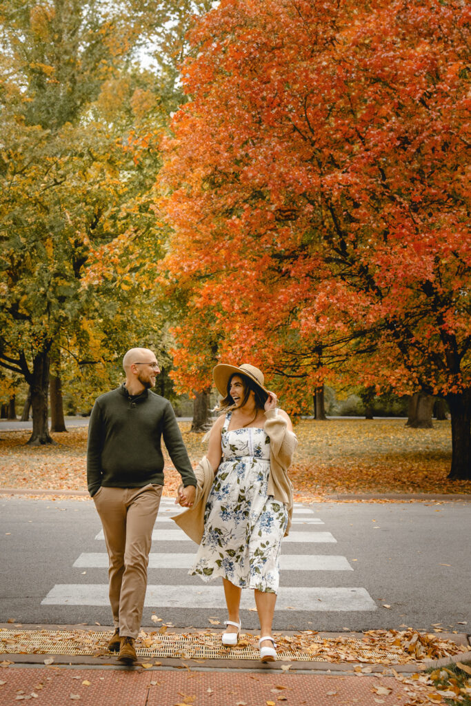 front shot of a couple holding hands and looking lovingly at each other as they cross a crosswalk towards the camera against vibrant fall park trees