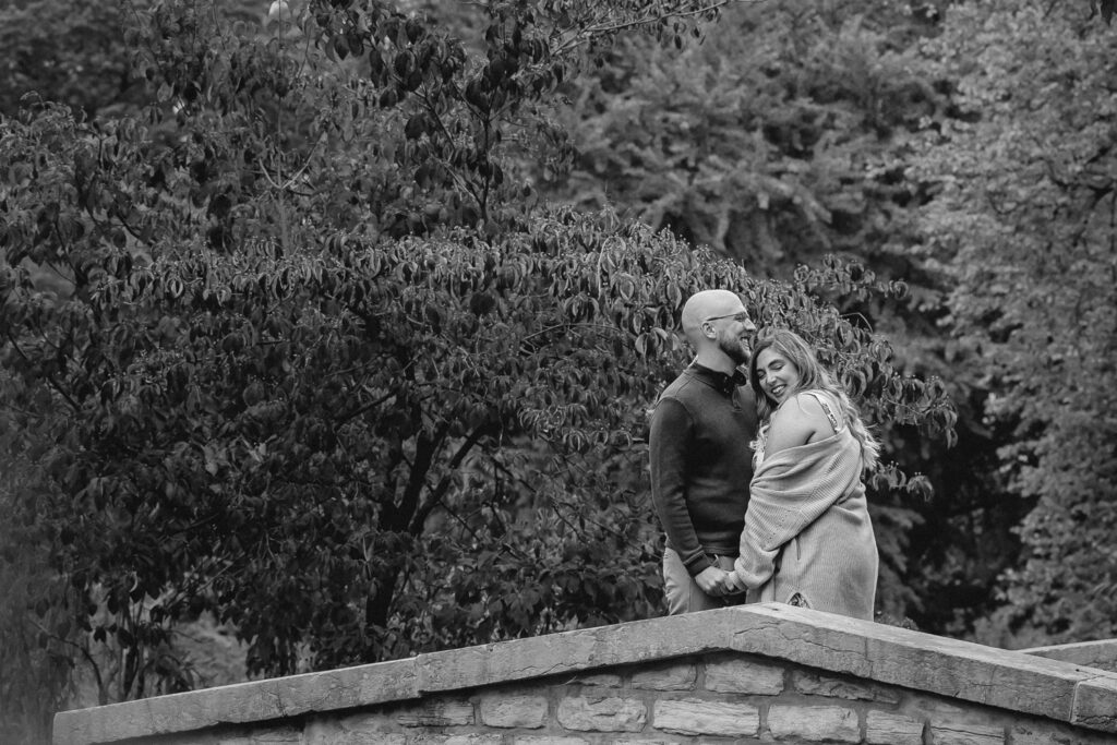 black and white of couple on a bridge, the boyfriend smiling off camera as his girlfriend smiles bashful towards the ground on his chest