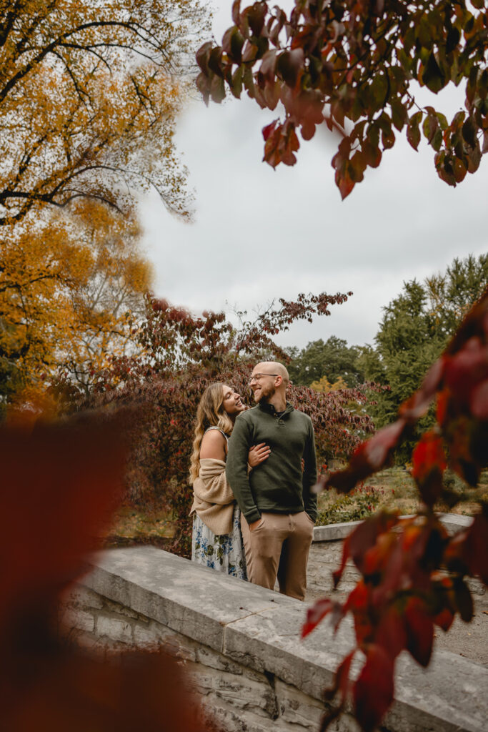 young man smiles back towards his girlfriend as she look up and hugs him from behind in colorful fall scene