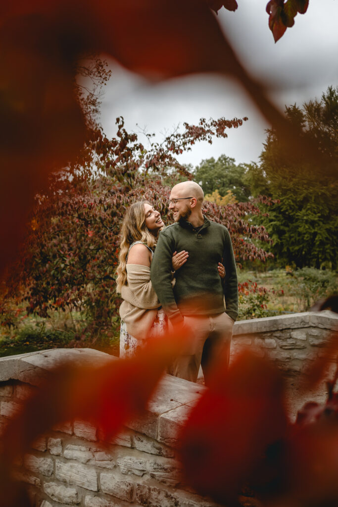young man smiles back towards his girlfriend as she look up and hugs him from behind between a frame blurry red leaves in the foreground