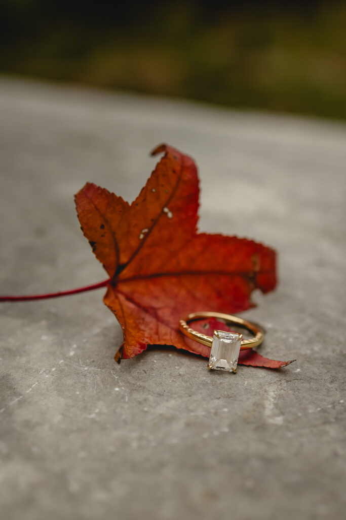 a gold engagement ring a large rectangular diamond sits on a red fall leaf sits perched on a stone railing