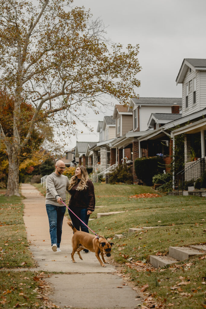front shot of couple walking their dog towards the camera down the sidewalk of a classic St. Louis South City-style neighborhood  with small unique brick homes