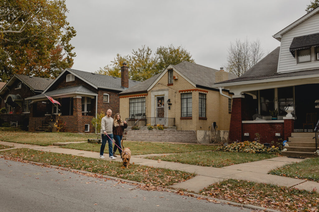 side shot of couple walking their dog on a leash on the sidewalk of a classic St. Louis South City-style neighborhood  with small unique brick homes