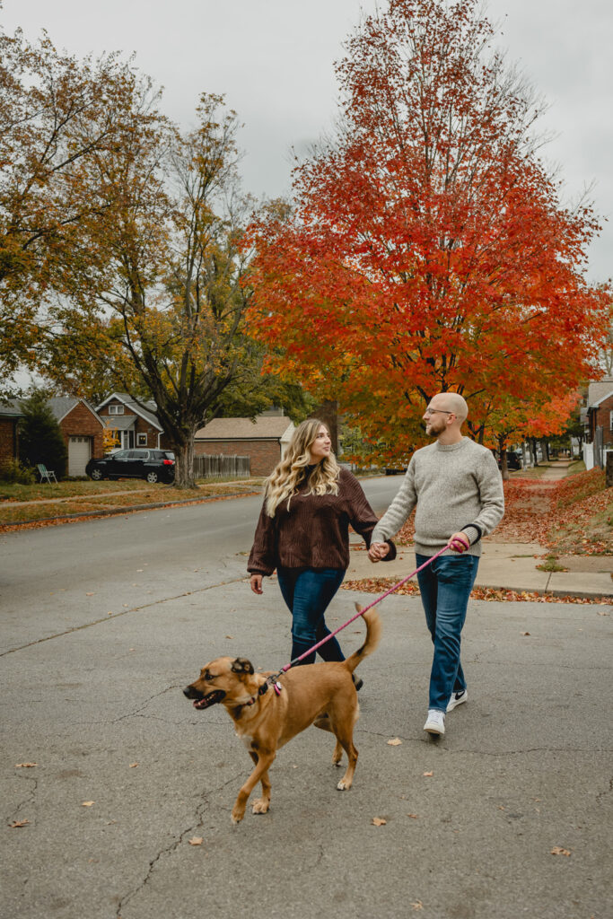 young couple smiles in casual sweaters walk across the street with their dog in a fall neighborhood 