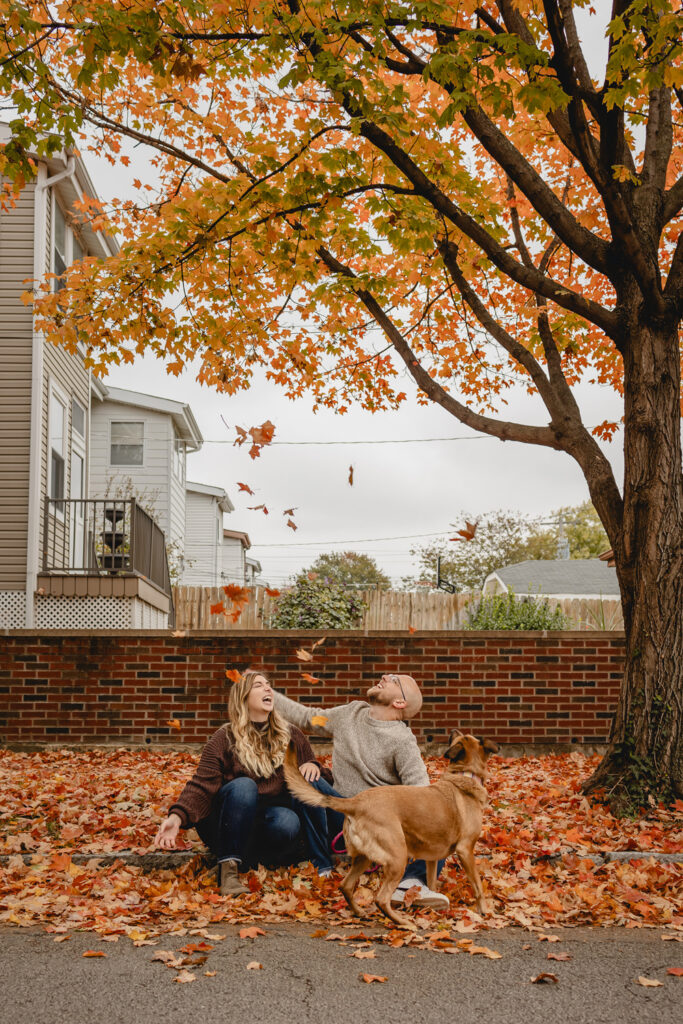 young couple sitting on a leaf covered curb as they try to time throwing leaves up in the air simultaneously