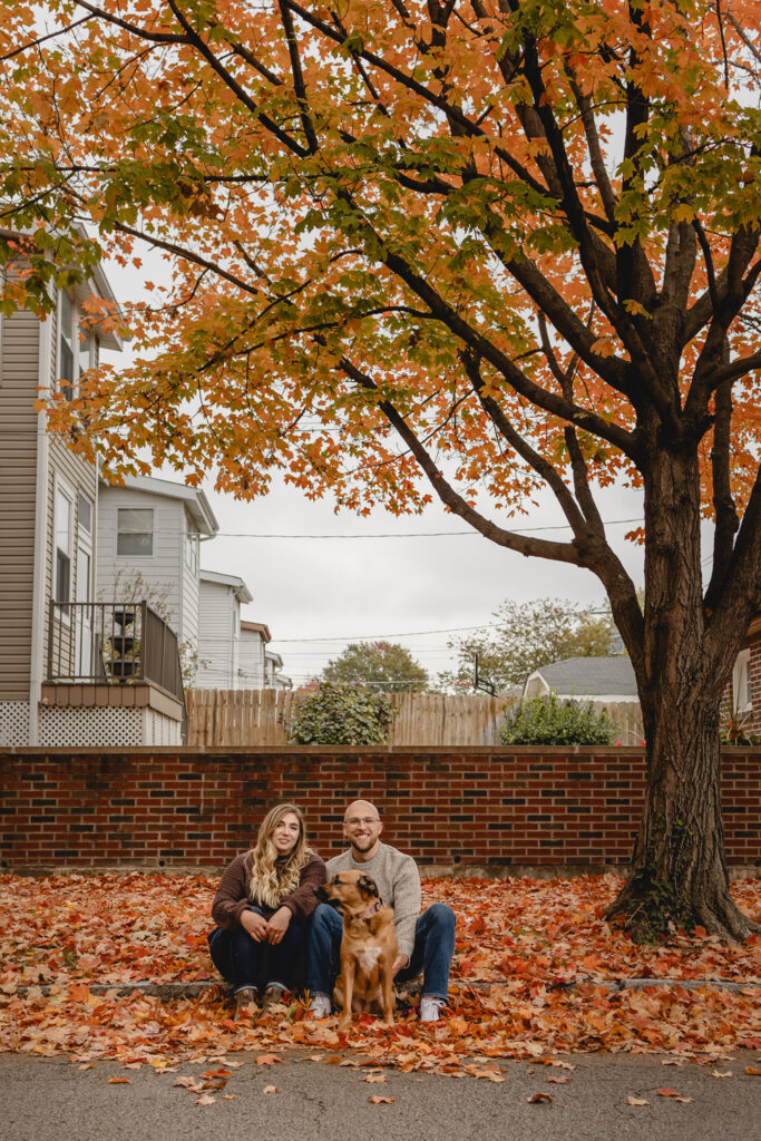 fall leaves cover a brick wall lined sidewalk in a neighborhood and a young couple sits centered in the middle with their dog.