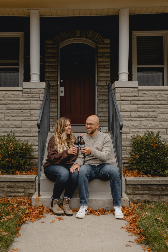 young couple sitting on short front porch steps of a limestone house smiling as they cheers bottles of local zwickel beer