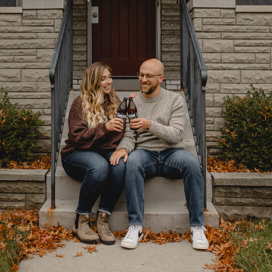 young couple sitting on short front porch steps of a limestone house smiling as they cheers bottles of local zwickel beer