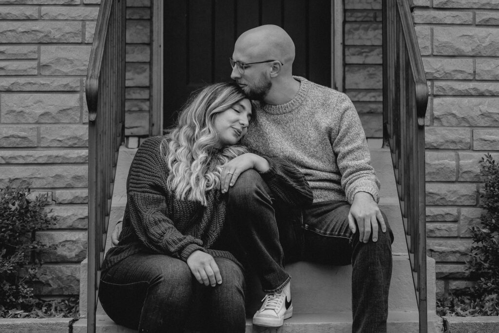 black and white shot of young woman peacfully resting on her boyfriends knee as he rest his head on hers while sitting on porch steps