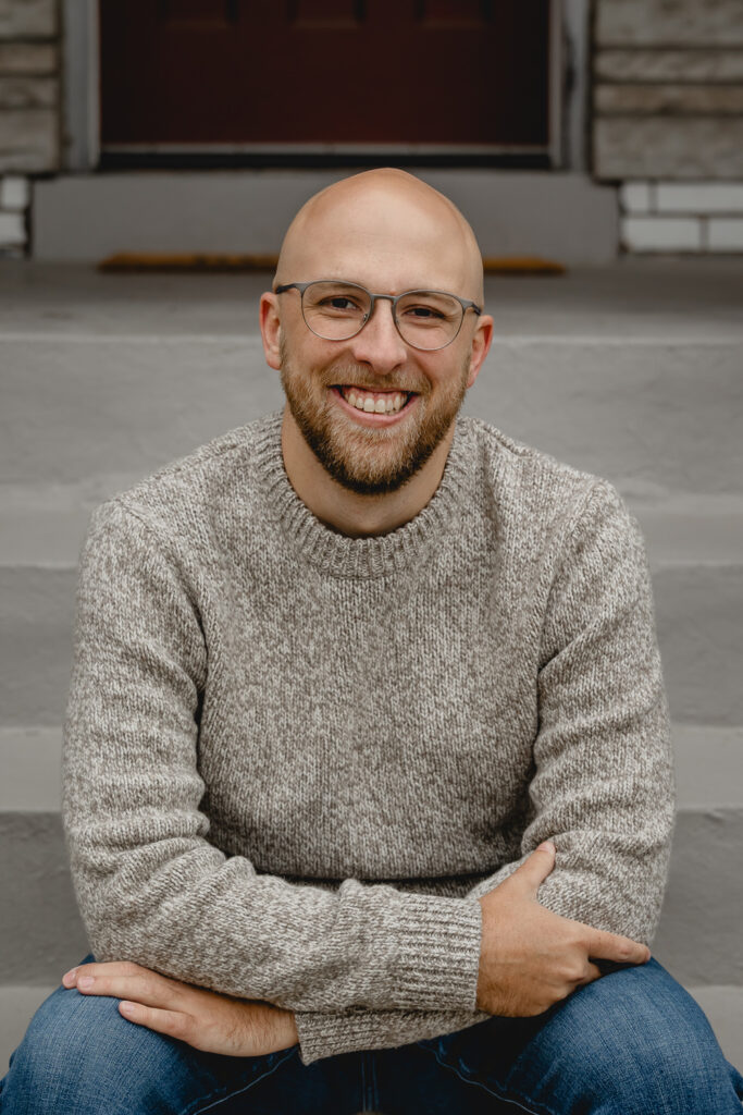 mid-body portrait of a young bald man with trimmed beard in a chunky gray sweater smiling to camera as he leans his arms against his knees