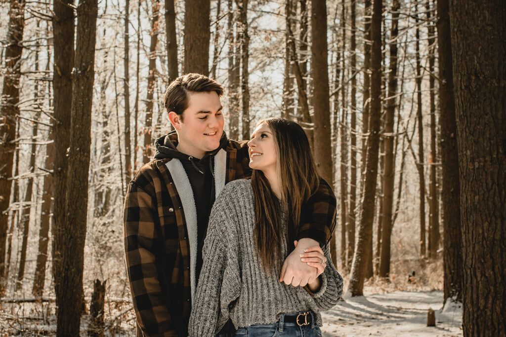 young women leans against her boyfriend and smiles back up his way in front of many tall snow covered tree trunks