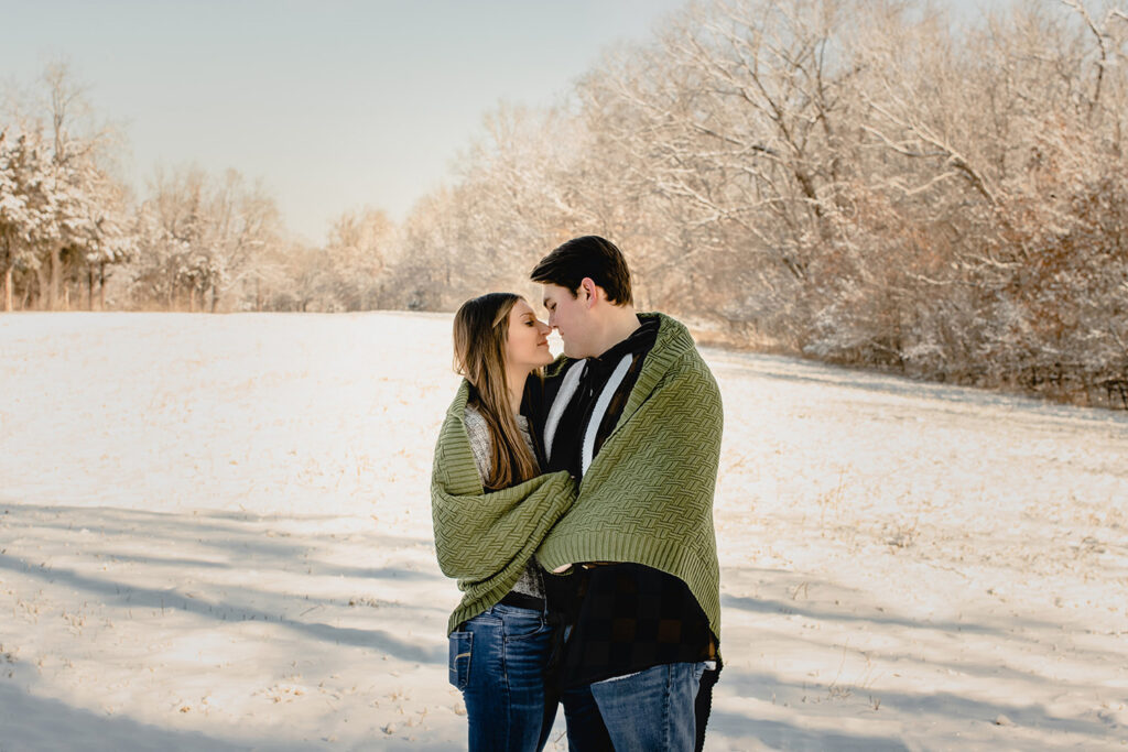 a young couple stand wrapped in a small green blanket and nuzzle noses in front of a snow-covered meadow