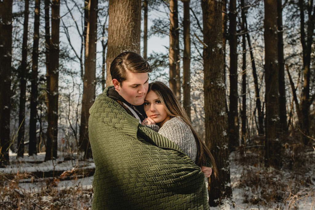 young man wraps his girlfriend in tight with a blanket as she looks serenely to the camera