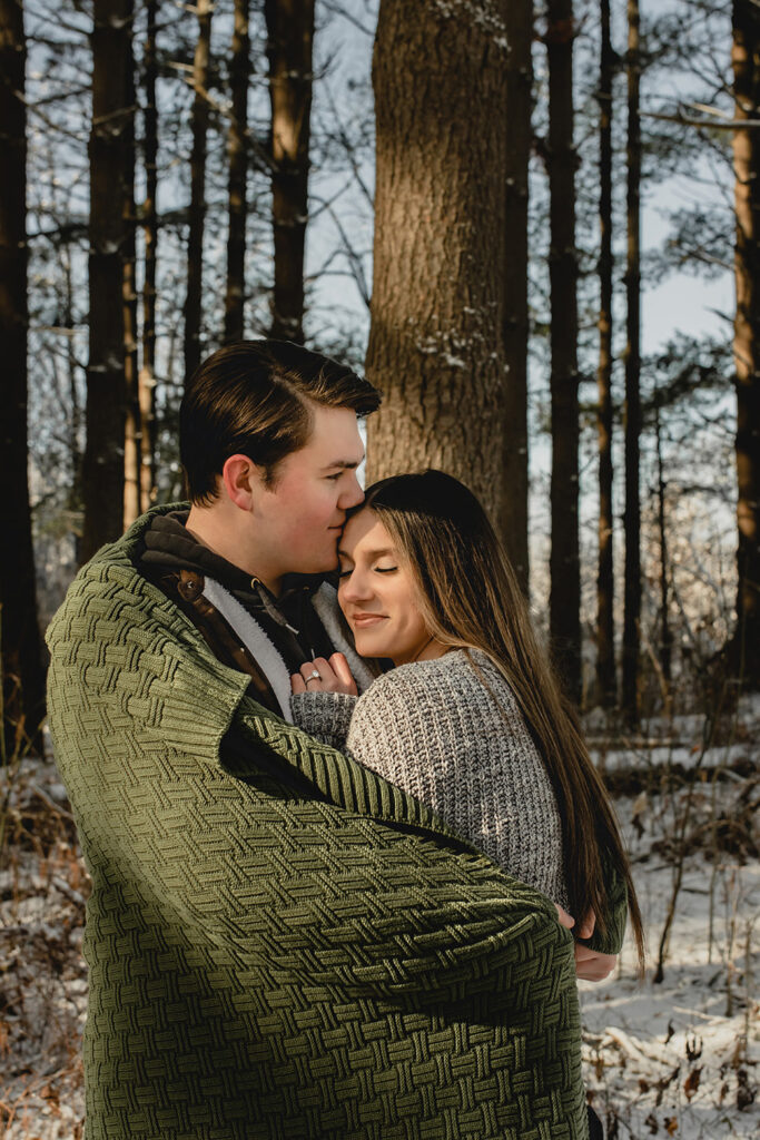 young man kisses his girlfriends forehead she she leans into his chest  in a winter forest