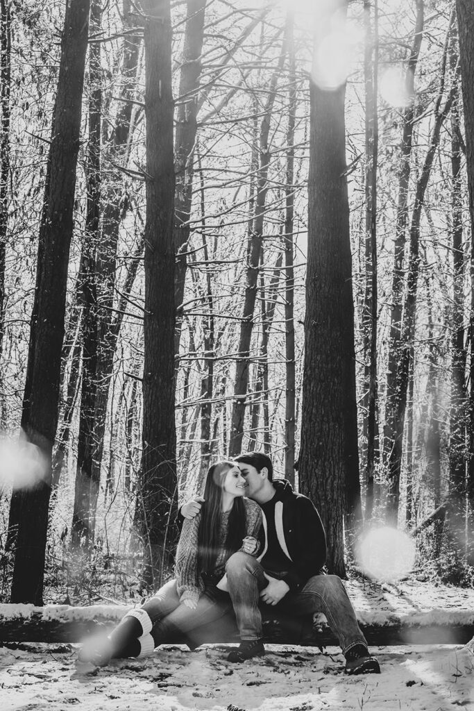 black and white candid of young couple sitting on a snow-covered log with snowflakes blurring parts of the image