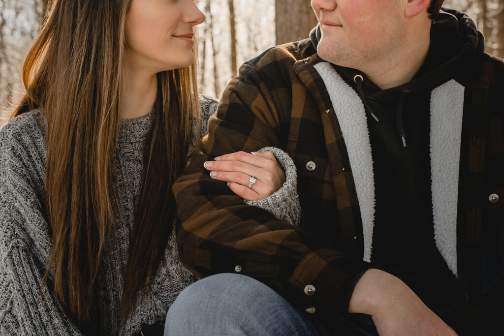 tight crop of a young couple smiling towards each other with arms wrapped to showcase an engagement ring