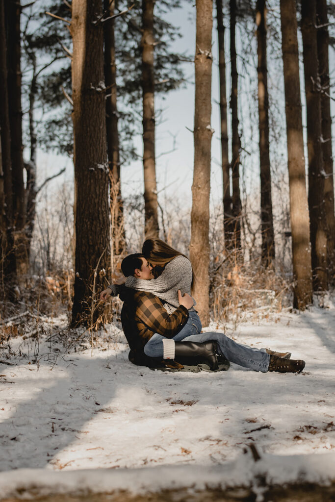 far shot of a woman sitting on her boyfriends lap with foreheads together on the snow-covered ground